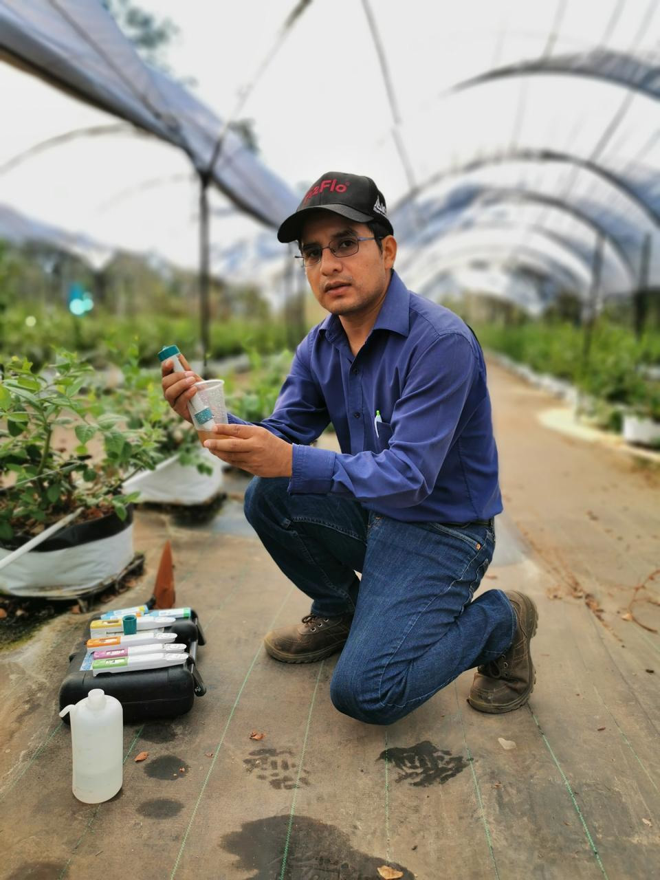 Mateo Martínez crouches in a greenhouse, performing agricultural analysis with a sample container and a testing kit.