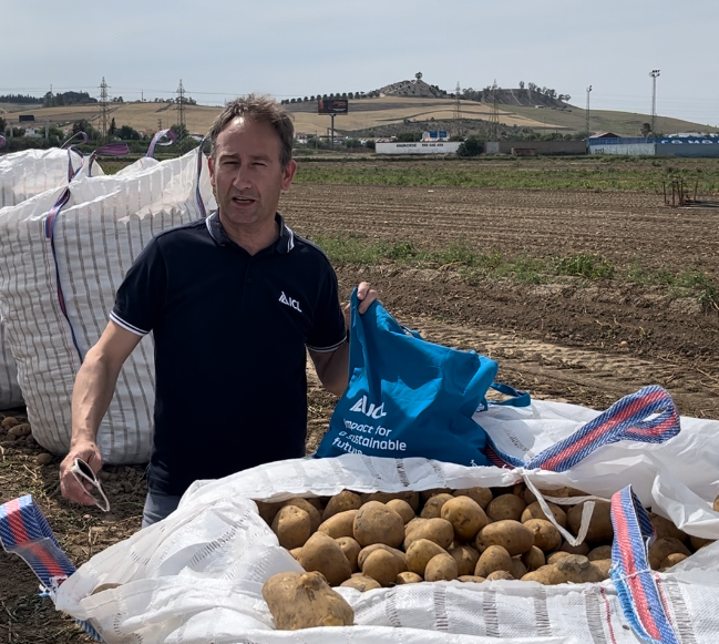 Manu Moreno stands in a field next to large sacks filled with freshly harvested potatoes, holding a blue tote bag.