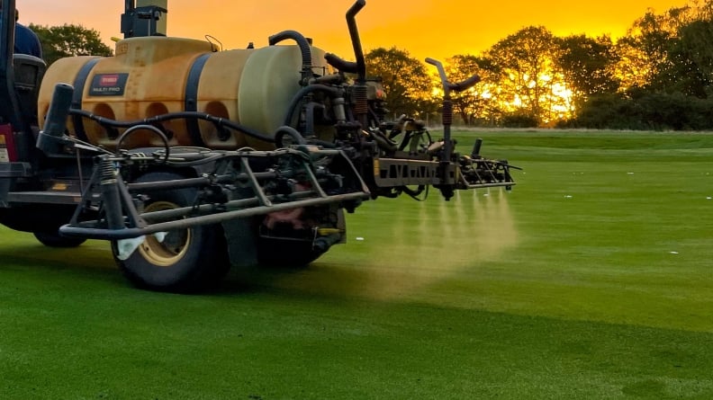 Sprayer applying a liquid treatment to a golf green during routine turf management at sunrise.