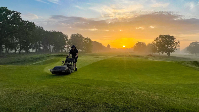 Greenkeeper mowing a golf green with a pedestrian mower in early morning light.