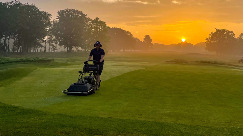 Greenkeeper mowing a closely maintained golf green with a pedestrian mower at sunrise.